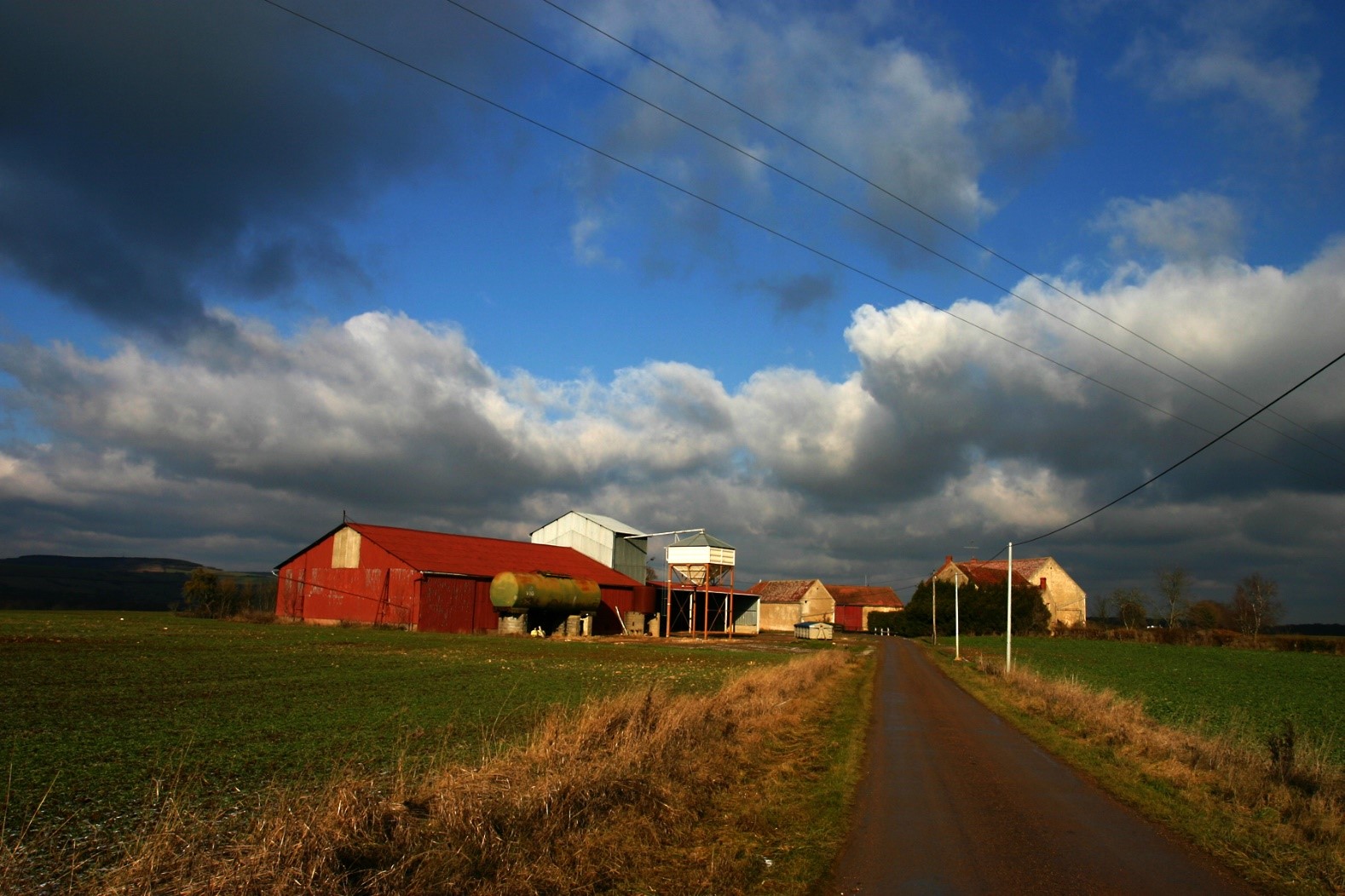 Visuel d'une route de campagne avec la vue d'une ferme agricole et des nuages très bas sous un ciel très bleu